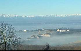 Weitläufiges Tal mit Nebel, vereinzelten Gebäuden und schneebedeckter Bergkette im Hintergrund.